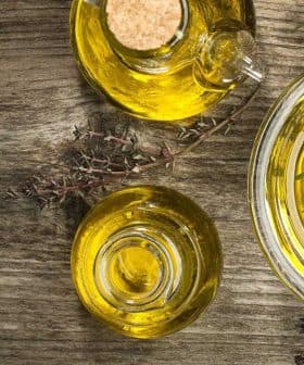 Glass bowl and bottle containing olive oil with herbs on a wooden surface. - Olive Oil Times