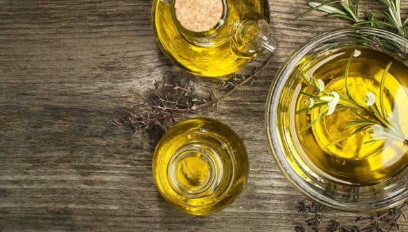 Glass bowl and bottle containing olive oil with herbs on a wooden surface. - Olive Oil Times