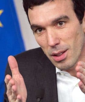 Man in a suit gesturing with hands during a discussion, European Union flag visible in the background. - Olive Oil Times