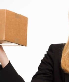 A woman in a black blazer holding a cardboard package with marketplace branding. - Olive Oil Times