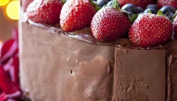 Chocolate cake topped with strawberries and blueberries, surrounded by rose petals. - Olive Oil Times