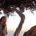 Woman with curly hair sitting on a branch of an olive tree during sunset. - Olive Oil Times