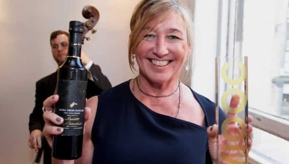 Woman in a navy dress holding a bottle of wine and a tall glass at an indoor event. - Olive Oil Times