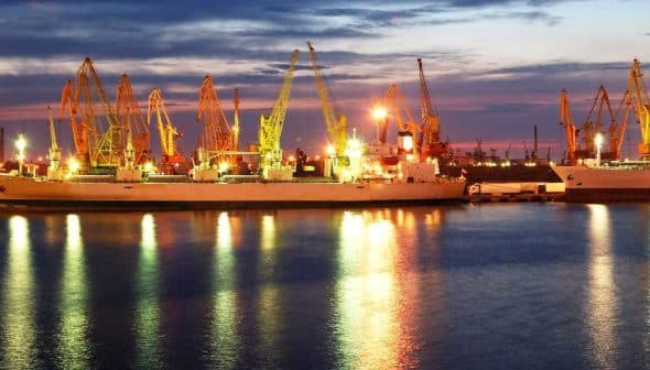Cargo ships docked at a port with cranes illuminated by lights during twilight hours. - Olive Oil Times