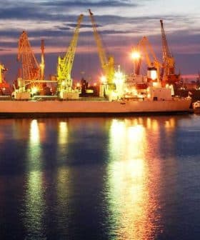 Cargo ships docked at a port with cranes illuminated at twilight. - Olive Oil Times