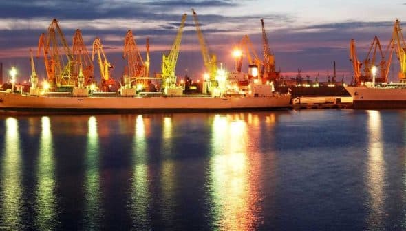 Cargo ships docked at a port with cranes illuminated at twilight. - Olive Oil Times