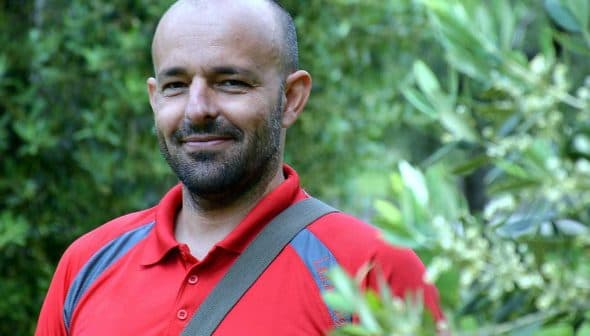 Man wearing a red polo shirt standing among olive trees with a slight smile. - Olive Oil Times
