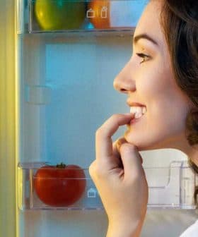 A woman with dark hair smiling while looking into an open refrigerator filled with food items. - Olive Oil Times