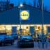 Exterior view of a Lidl supermarket with a lit sign and parked cars in front during dusk. - Olive Oil Times