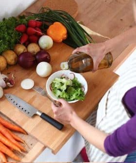 Individual pouring oil over a bowl of salad with various fresh vegetables on a wooden countertop. - Olive Oil Times