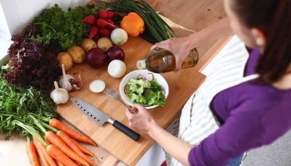 Individual pouring oil over a bowl of salad with various fresh vegetables on a wooden countertop. - Olive Oil Times