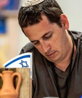 Man wearing a kippah writing at a table with American and Israeli flags in the background. - Olive Oil Times
