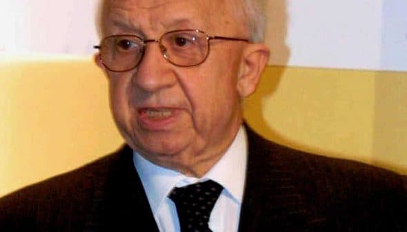 Elderly man wearing glasses and a dark suit speaking during an event. - Olive Oil Times