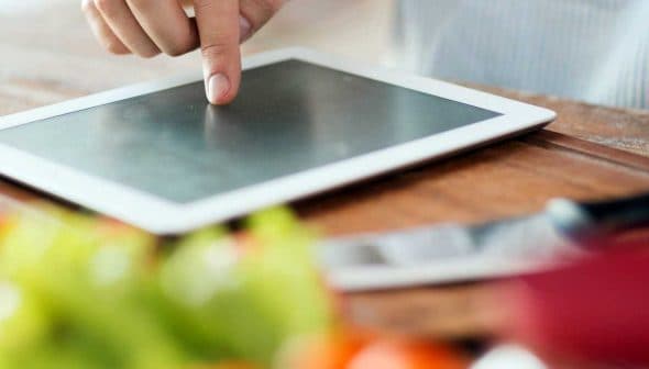 A person pointing at a tablet screen while preparing food in a kitchen setting. - Olive Oil Times