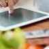 A person pointing at a tablet screen while preparing food in a kitchen setting. - Olive Oil Times