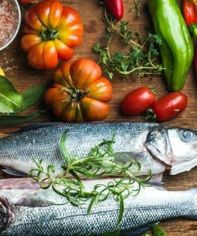 Two whole fish placed on a wooden cutting board surrounded by various vegetables and herbs. - Olive Oil Times