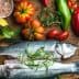 Two whole fish placed on a wooden cutting board surrounded by various vegetables and herbs. - Olive Oil Times