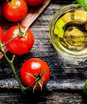 Fresh red tomatoes, green basil leaves, and a small glass of olive oil on a wooden surface. - Olive Oil Times