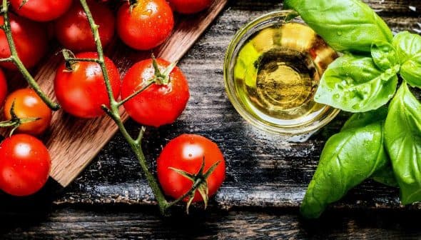 Fresh red tomatoes, green basil leaves, and a small glass of olive oil on a wooden surface. - Olive Oil Times