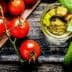 Fresh red tomatoes, green basil leaves, and a small glass of olive oil on a wooden surface. - Olive Oil Times
