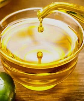 A glass bowl filled with olive oil with a stream pouring from a vessel and olives nearby. - Olive Oil Times