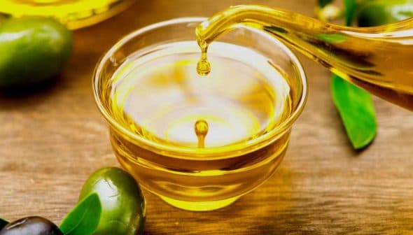 A glass bowl filled with olive oil with a stream pouring from a vessel and olives nearby. - Olive Oil Times
