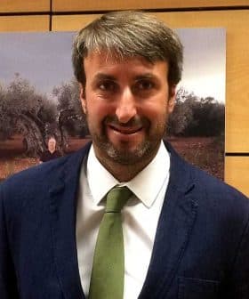 Man wearing a dark suit and a green tie, standing in front of a background featuring olive trees. - Olive Oil Times