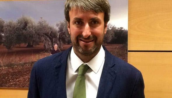 Man wearing a dark suit and a green tie, standing in front of a background featuring olive trees. - Olive Oil Times