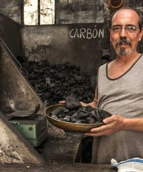 Man in a tank top holding a bowl of charcoal in a dimly lit room with charcoal piles. - Olive Oil Times