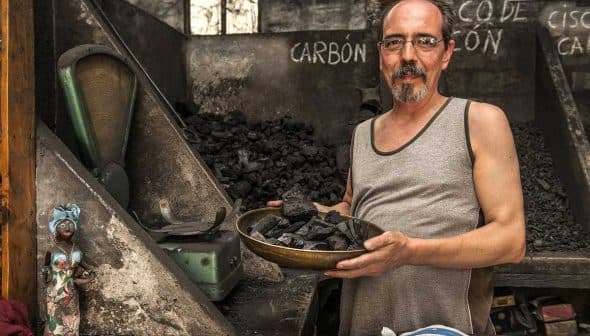 Man in a tank top holding a bowl of charcoal in a dimly lit room with charcoal piles. - Olive Oil Times