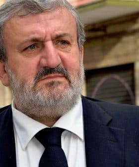 A man with a gray beard wearing a dark suit and tie, looking serious outdoors. - Olive Oil Times