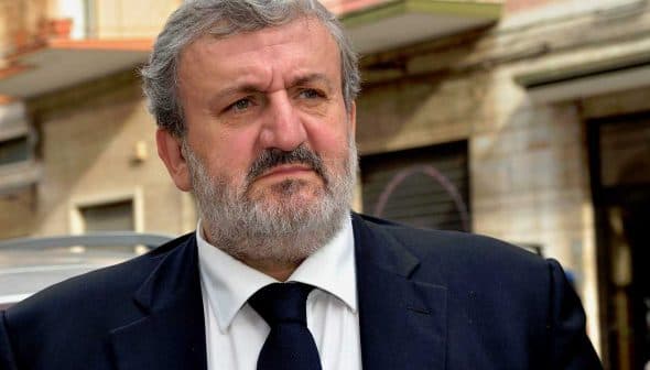 A man with a gray beard wearing a dark suit and tie, looking serious outdoors. - Olive Oil Times