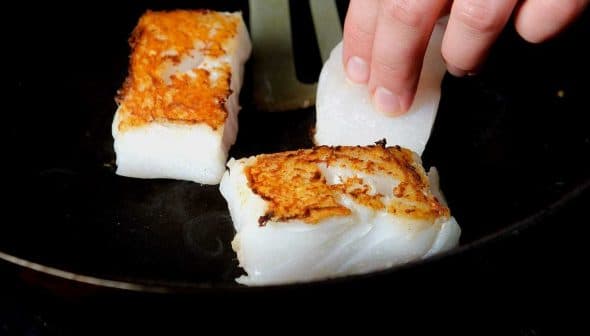 Person placing fish fillets into a skillet while cooking on a stovetop. - Olive Oil Times