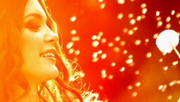 A woman smiling while holding a dandelion seed head in her hand, with a blurred background. - Olive Oil Times