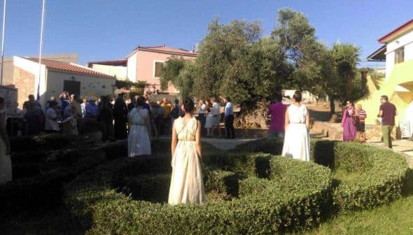 A gathering of people outdoors with dancers in white costumes in a garden setting. - Olive Oil Times