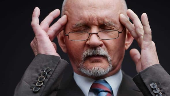 A man with gray hair and glasses holding his head with both hands, appearing deep in thought. - Olive Oil Times