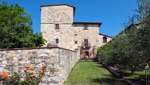A stone building featuring a tower and surrounded by a garden with roses and greenery. - Olive Oil Times