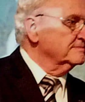 An older man in a black suit and glasses speaking on stage at an event. - Olive Oil Times