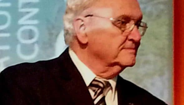 An older man in a black suit and glasses speaking on stage at an event. - Olive Oil Times