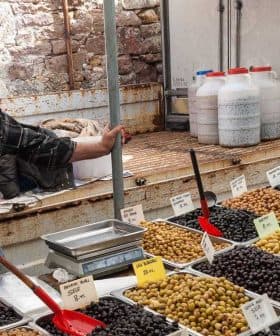 A vendor stands beside trays of various olives at a market stall, with labels indicating prices. - Olive Oil Times