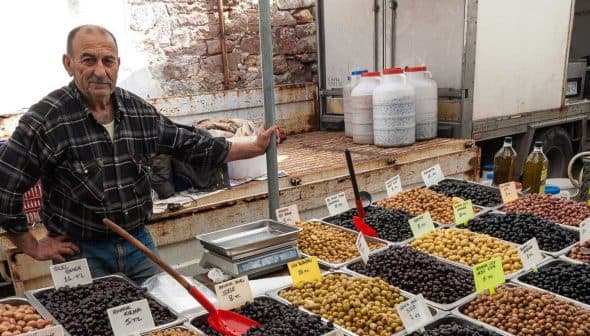 A vendor stands beside trays of various olives at a market stall, with labels indicating prices. - Olive Oil Times