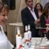 A woman smiling while serving olive oil at a tasting event with attendees in the background. - Olive Oil Times