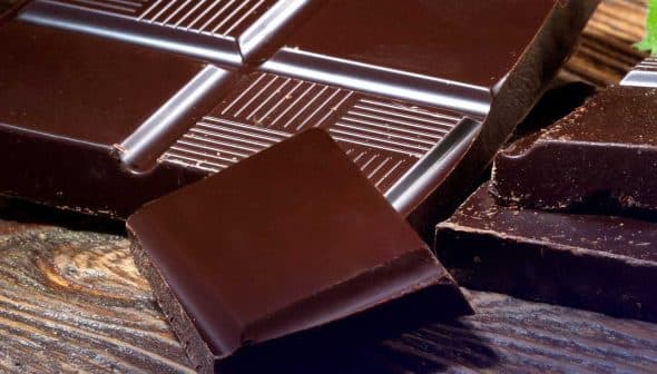 A dark chocolate bar with several pieces broken off, placed on a wooden surface. - Olive Oil Times