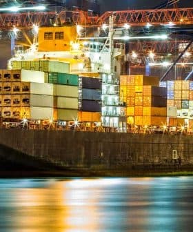 Large cargo ship docked at a port, loaded with colorful shipping containers under night lights. - Olive Oil Times