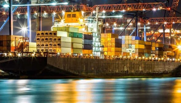 Large cargo ship docked at a port, loaded with colorful shipping containers under night lights. - Olive Oil Times