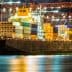 Large cargo ship docked at a port, loaded with colorful shipping containers under night lights. - Olive Oil Times