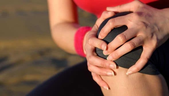 Individual with a red shirt holding their knee with both hands, indicating discomfort or pain. - Olive Oil Times