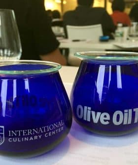 Two blue glass cups labeled 'International Culinary Center' and 'Olive Oil Times' on a table. - Olive Oil Times