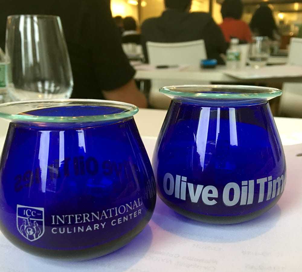 Two blue glass cups labeled 'International Culinary Center' and 'Olive Oil Times' on a table. - Olive Oil Times