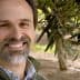 Man with a beard smiling in an olive grove with olive branches in the background. - Olive Oil Times